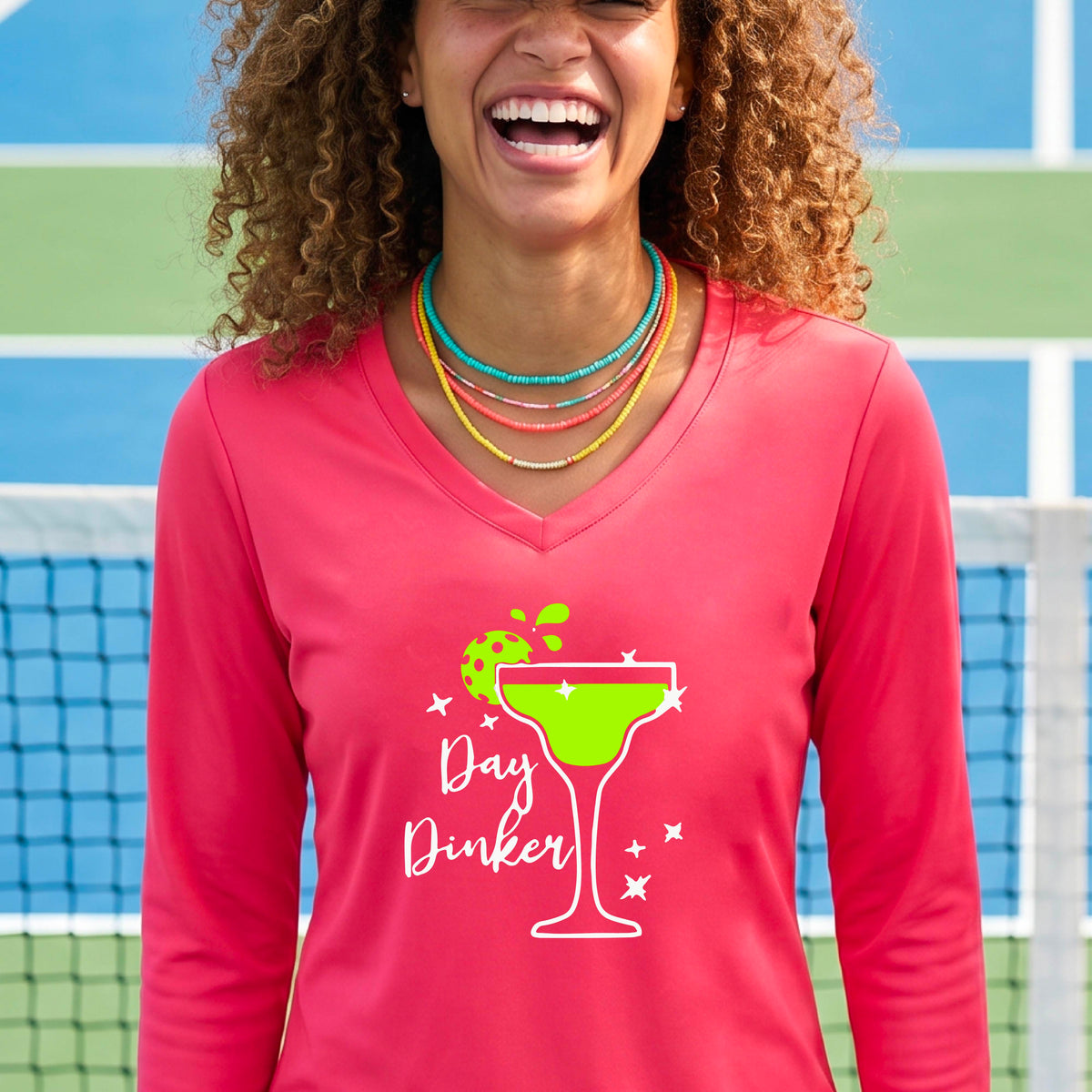 A woman wearing a coral pink  long-sleeve V-neck t-shirt with a 'Day Dinker' martini glass graphic in green and white with a pickleball lime garnish. 
