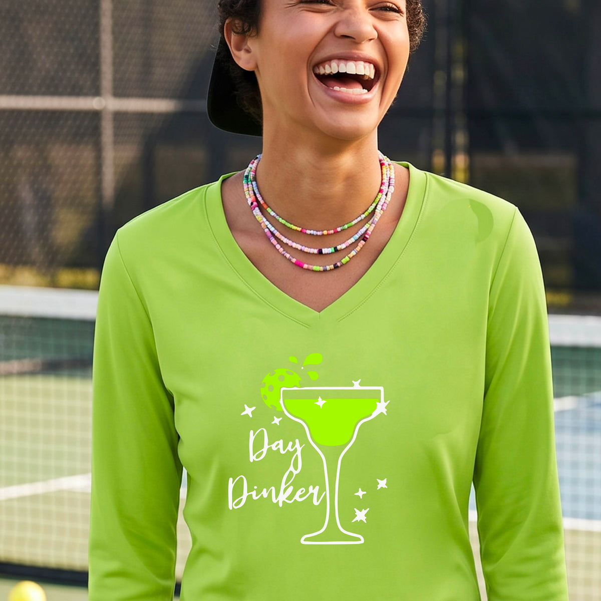 A woman wearing a lime green long-sleeve V-neck t-shirt with a 'Day Dinker' martini glass graphic in green and white with a pickleball lime garnish. 