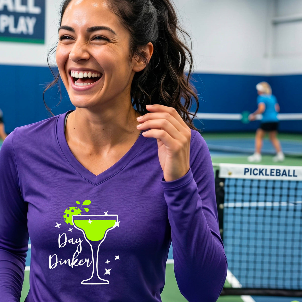 A woman wearing a purple long-sleeve V-neck t-shirt with a 'Day Dinker' martini glass graphic in green and white with a pickleball lime garnish. 