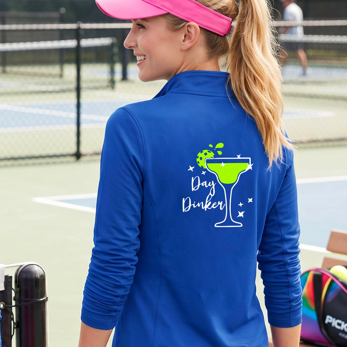 A woman on a pickleball court wearing a bright royal blue quarter-zip-up jacket, shown from the back. The back of the jacket features a white graphic margarita glass with a green pickleball lime garnish and the words 'Day Dinker' in white script, along with small white stars.