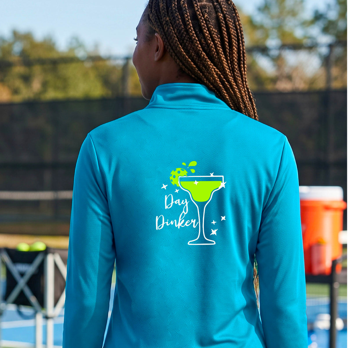 A woman on a pickleball court wearing a bright blue quarter-zip-up jacket, shown from the back. The back of the jacket features a white graphic margarita glass with a green pickleball lime garnish and the words 'Day Dinker' in white script, along with small white stars.