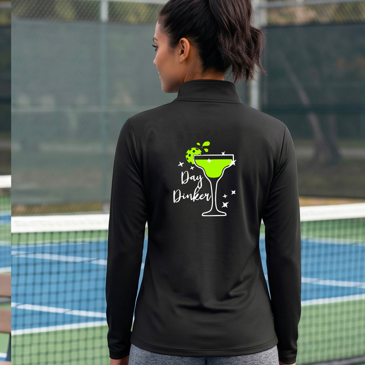A woman on a pickleball court wearing a black quarter-zip-up jacket, shown from the back. The back of the jacket features a white graphic margarita glass with a green pickleball lime garnish and the words 'Day Dinker' in white script, along with small white stars.