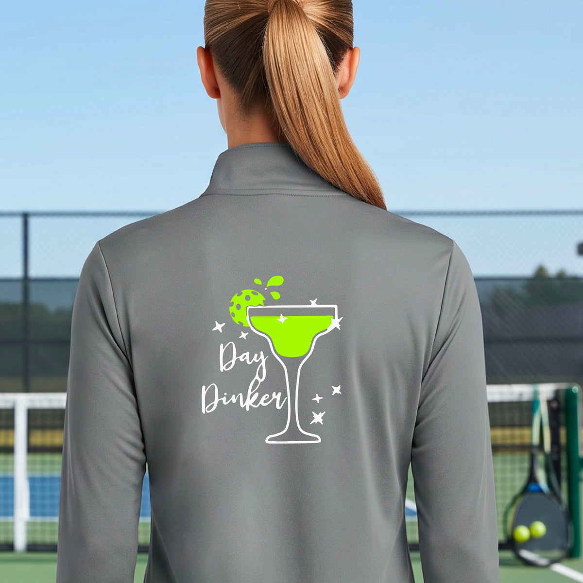 A woman on a pickleball court wearing a gray quarter-zip-up jacket, shown from the back. The back of the jacket features a white graphic margarita glass with a green pickleball lime garnish and the words 'Day Dinker' in white script, along with small white stars.