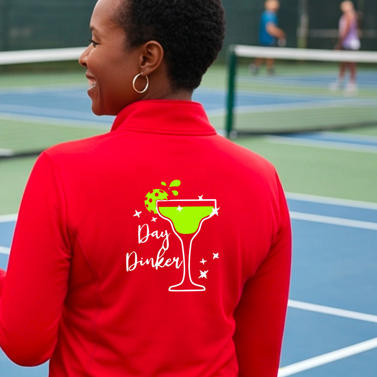 A woman on a pickleball court wearing a bright red quarter-zip-up jacket, shown from the back. The back of the jacket features a white graphic margarita glass with a green pickleball lime garnish and the words 'Day Dinker' in white script, along with small white stars.