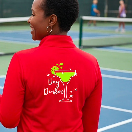 A woman on a pickleball court wearing a bright red quarter-zip-up jacket, shown from the back. The back of the jacket features a white graphic margarita glass with a green pickleball lime garnish and the words 'Day Dinker' in white script, along with small white stars.
