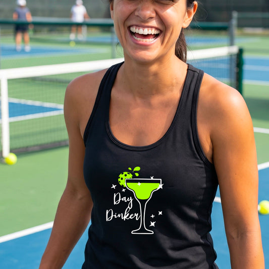 A woman on a pickleball court wearing a black racerback tank top with a 'Day Dinker' graphic featuring a green margarita glass, pickleball lime garnish and white script text.