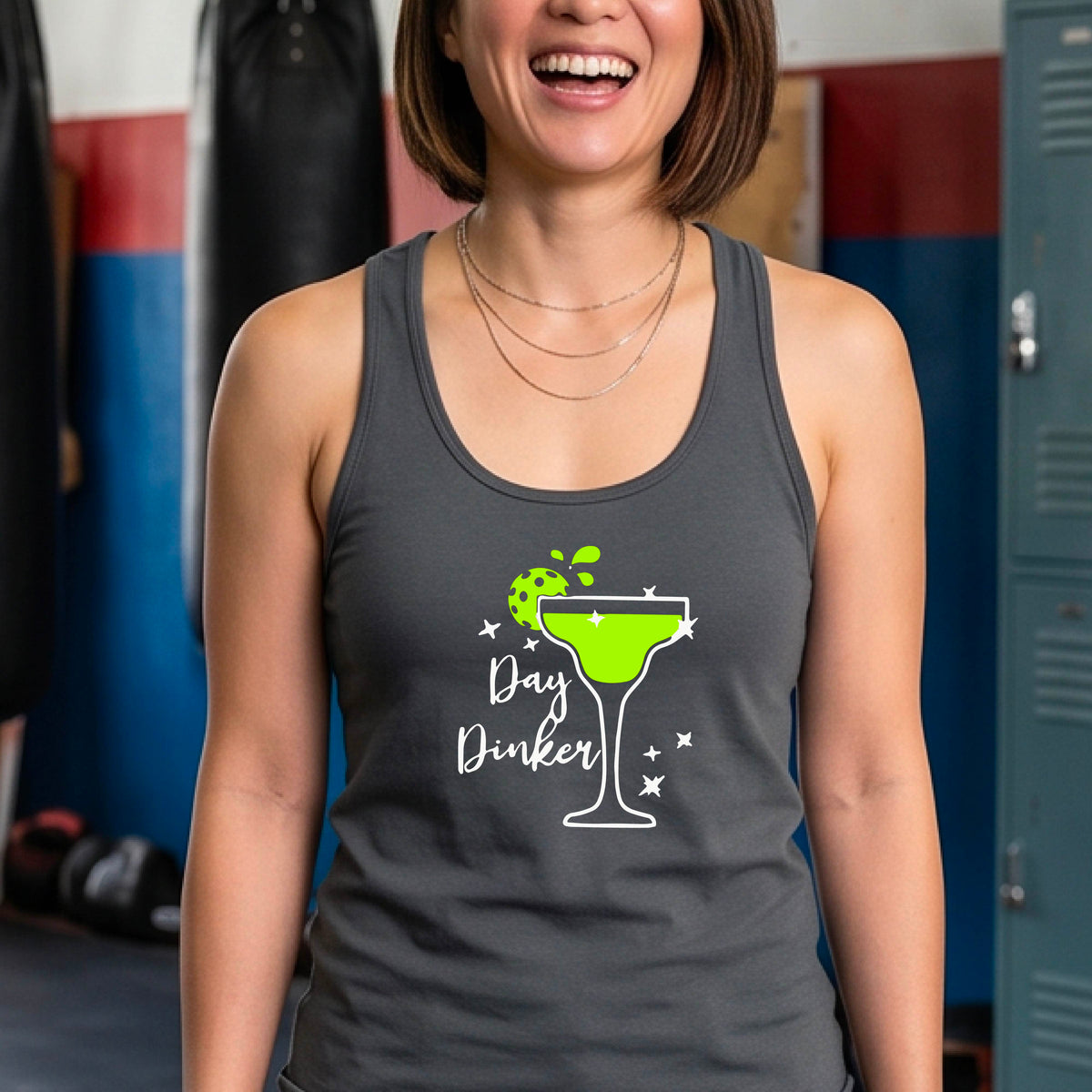A woman on a pickleball court wearing a dark gray racerback tank top with a 'Day Dinker' graphic featuring a green margarita glass, pickleball lime garnish and white script text.