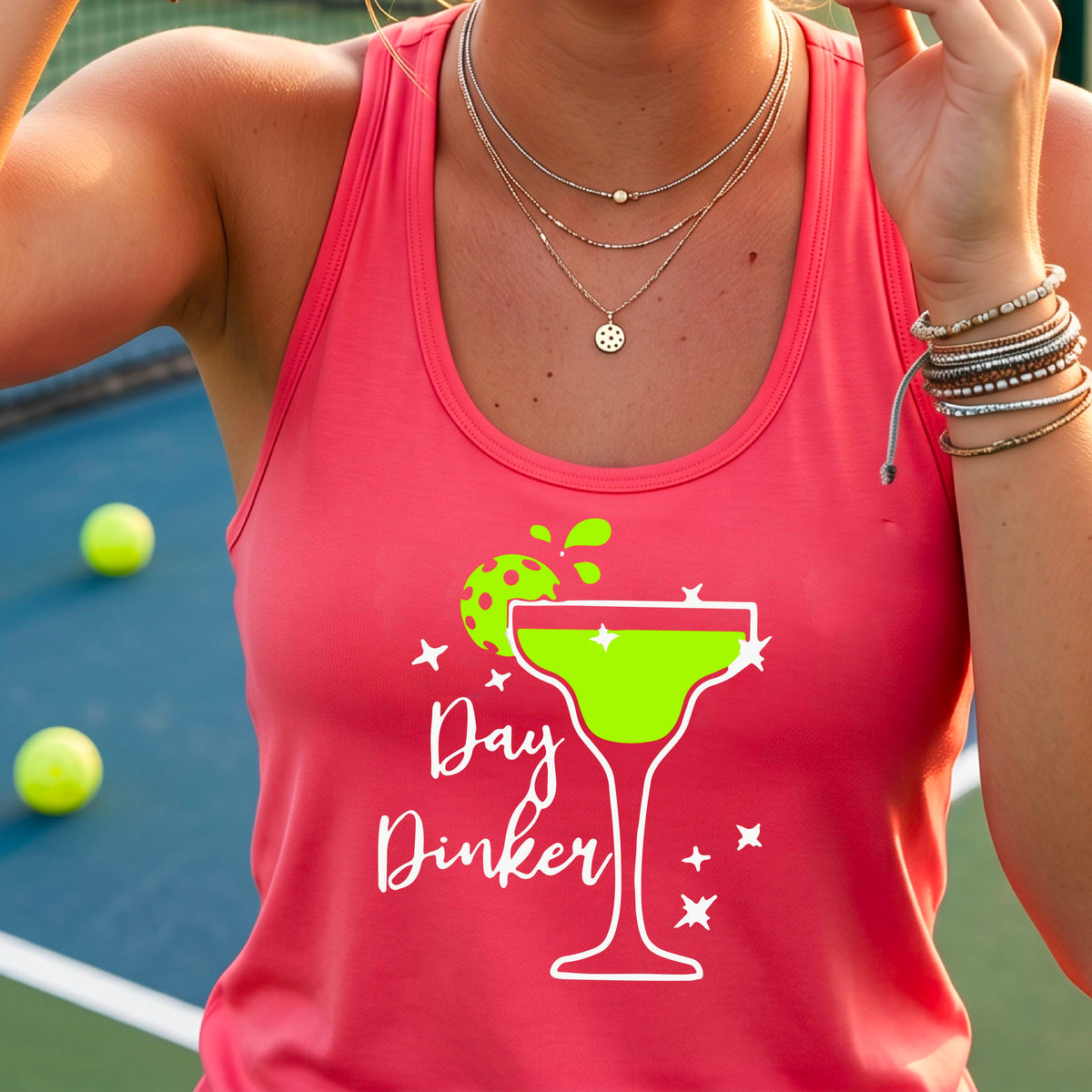 A woman on a pickleball court wearing a coral pink racerback tank top with a 'Day Dinker' graphic featuring a green margarita glass, pickleball lime garnish and white script text.