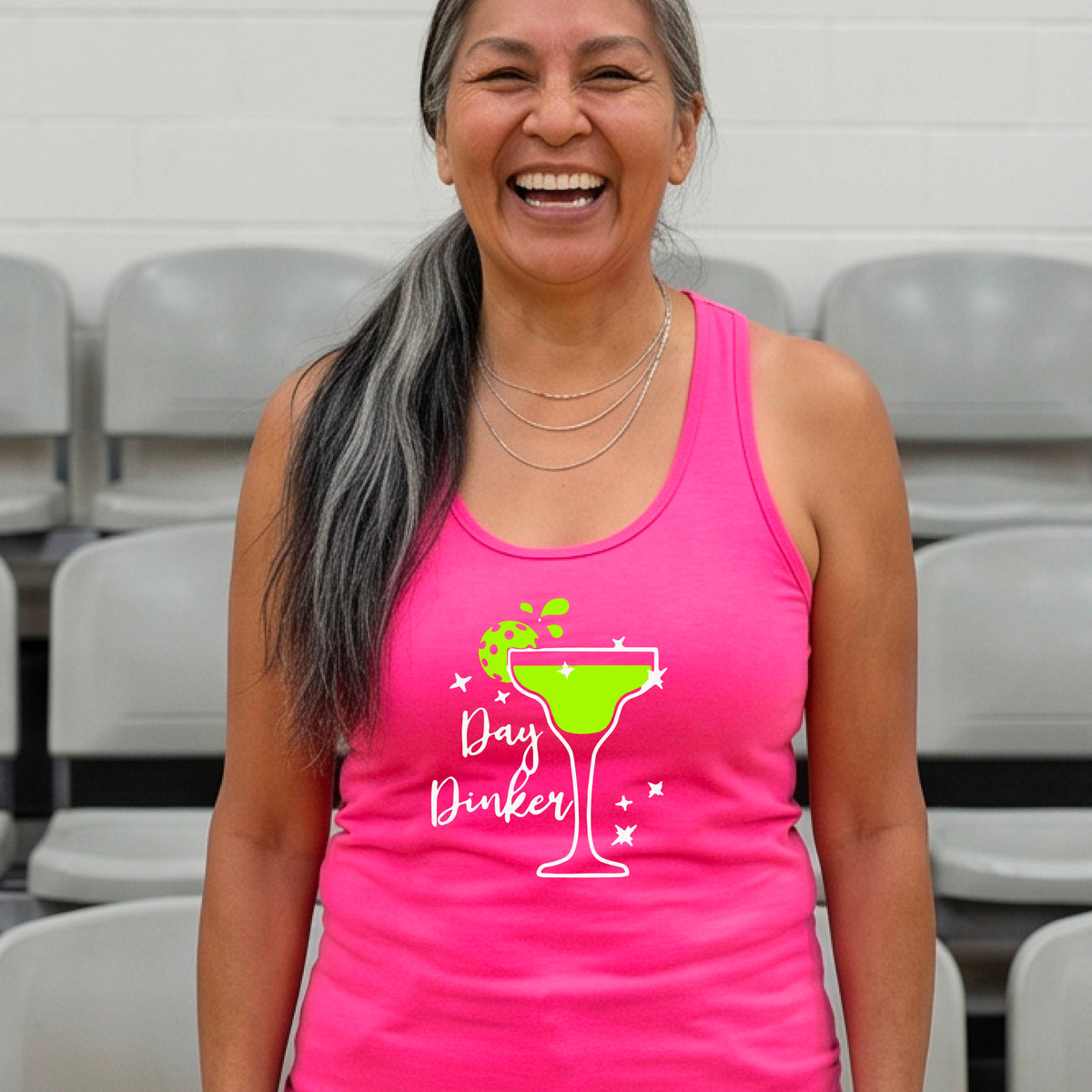 A woman on a pickleball court wearing a bright pink racerback tank top with a 'Day Dinker' graphic featuring a green margarita glass, pickleball lime garnish and white script text.