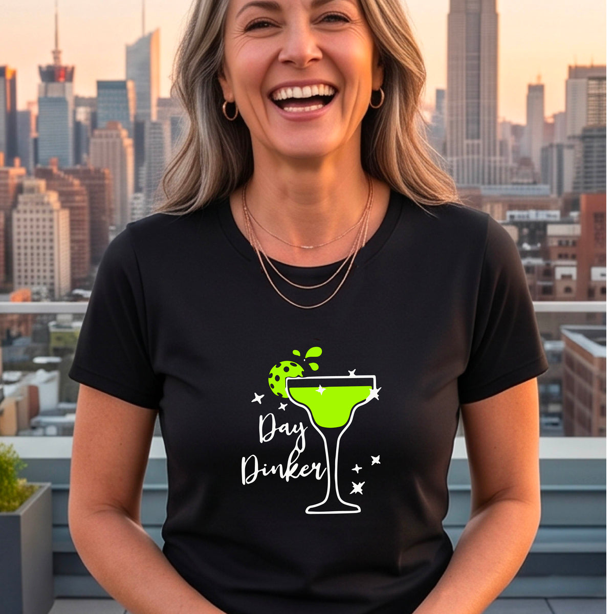 A smiling woman wearing a black graphic t-shirt with a green and white 'Day Dinker' cocktail design with a green pickleball lime garnish. 