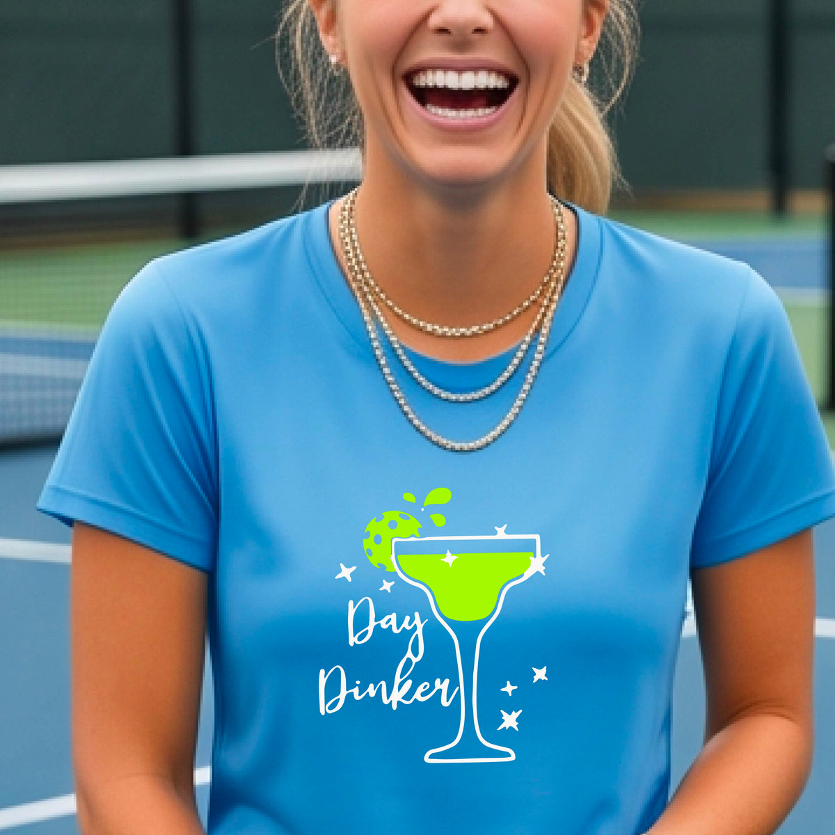 A woman wearing a bright blue t-shirt with a green and white 'Day Dinker' martini graphic with a lime pickleball garnish , standing on an outdoor pickleball court.