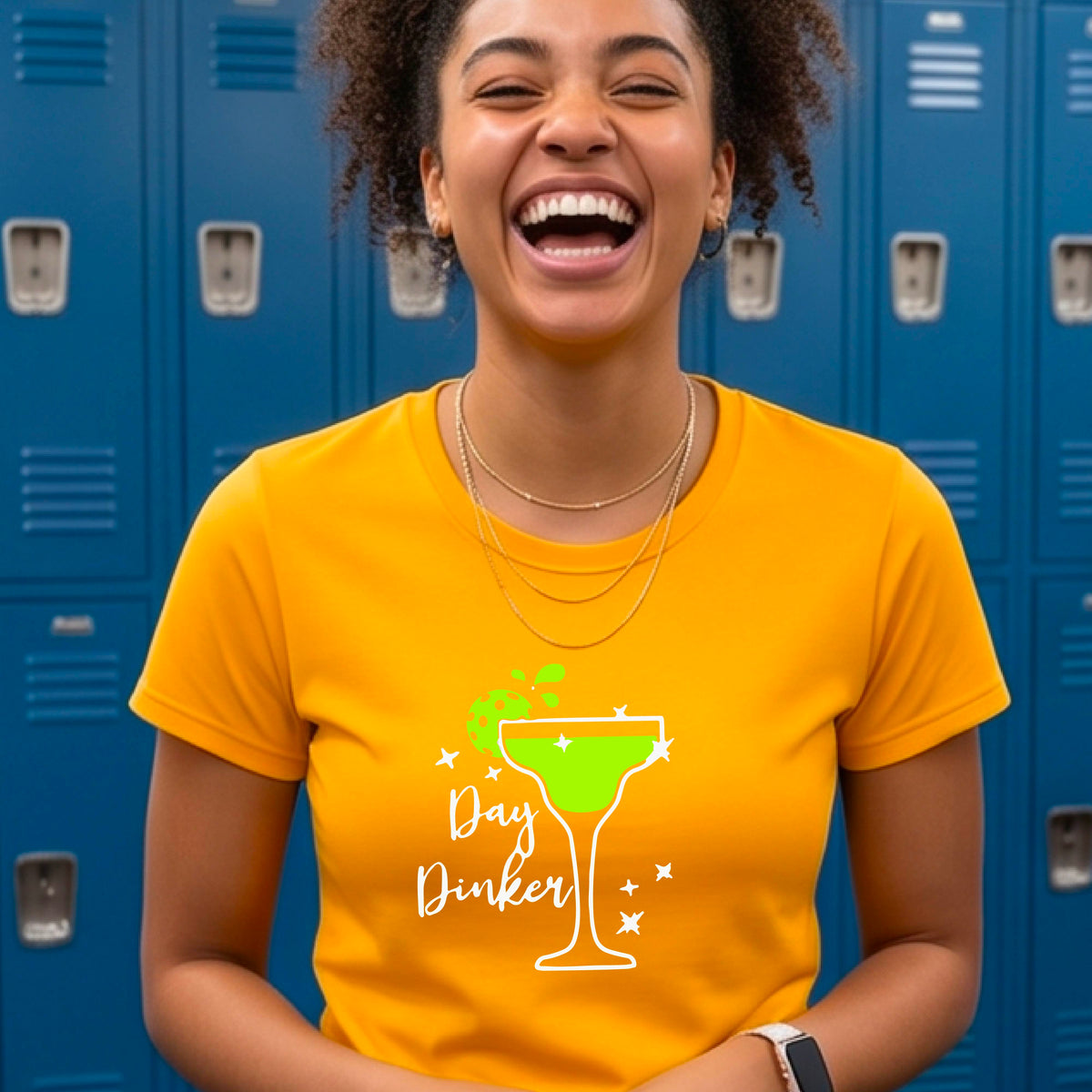A woman wearing a bright mustard yellow  t-shirt with a green and white 'Day Dinker' martini graphic with a lime pickleball garnish , standing on an outdoor pickleball court.