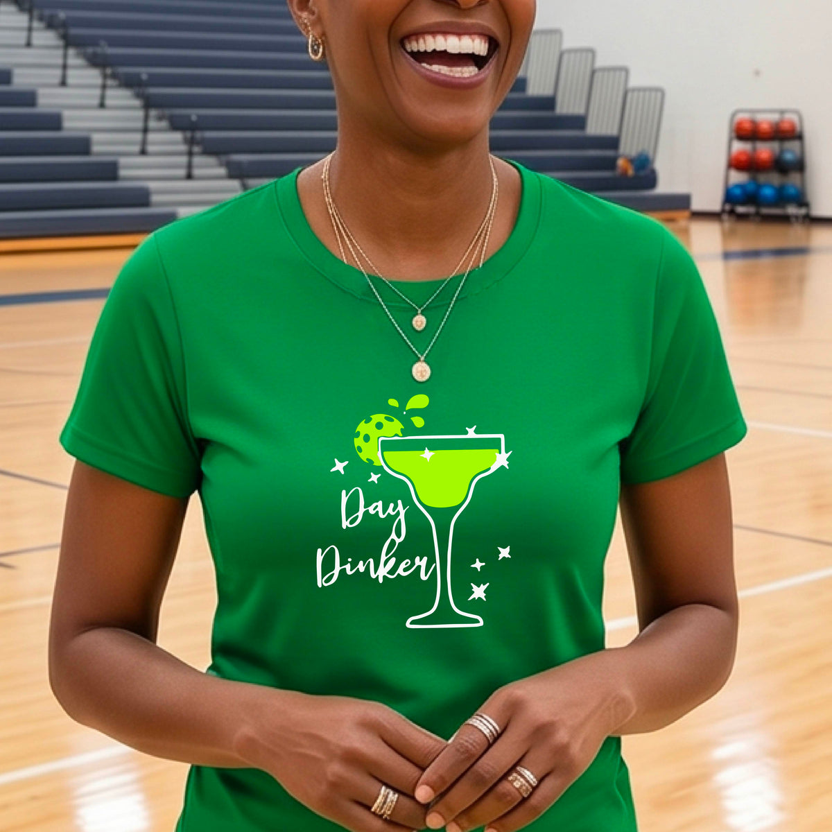 A woman wearing a bright green  t-shirt with a green and white 'Day Dinker' martini graphic with a lime pickleball garnish , standing on an outdoor pickleball court.