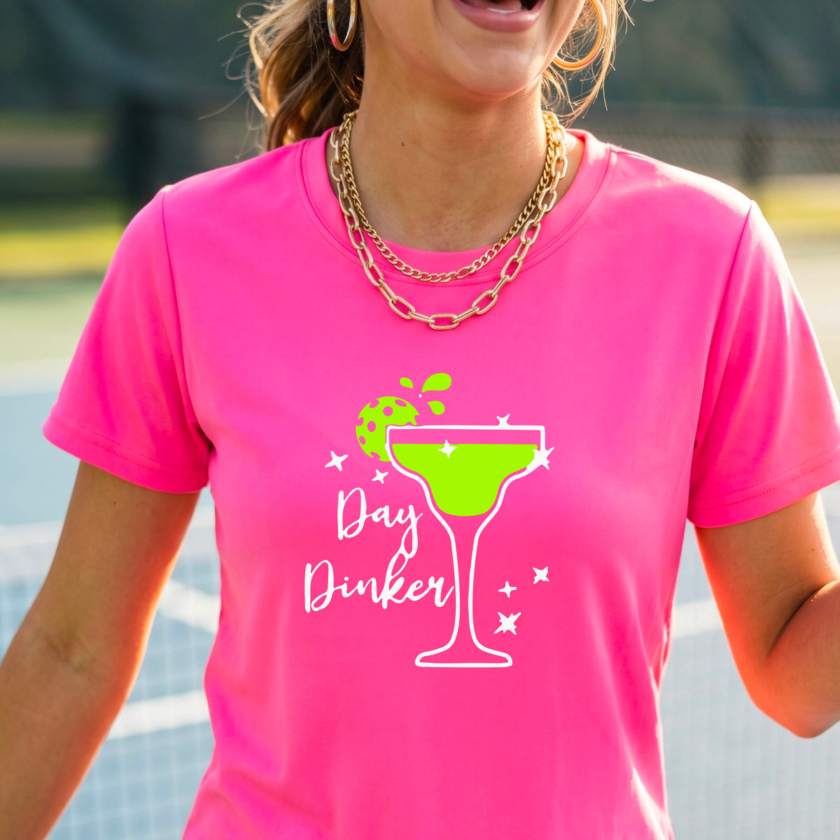 A woman wearing a bright pink t-shirt with a green and white 'Day Dinker' martini graphic with a lime pickleball garnish , standing on an outdoor pickleball court.