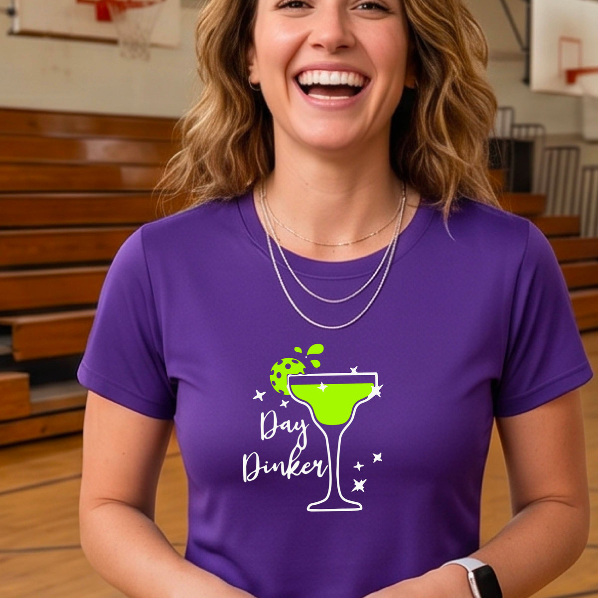 A woman wearing a purple t-shirt with a green and white 'Day Dinker' martini graphic with a lime pickleball garnish , standing on an outdoor pickleball court.