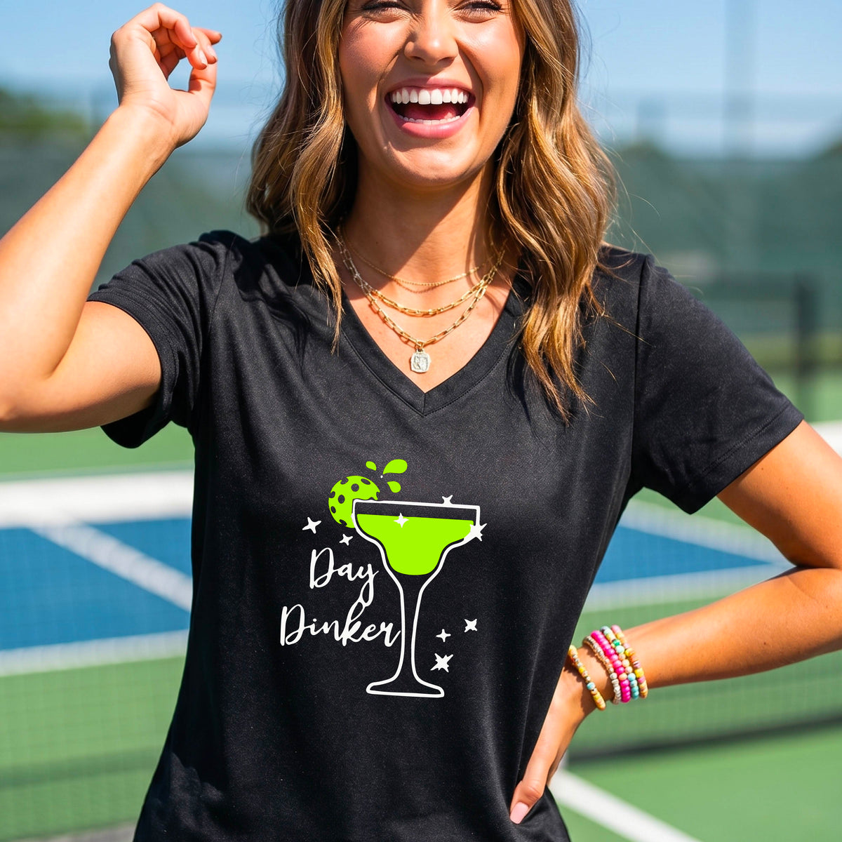 A woman wearing a black V-neck t-shirt with a 'Day Dinker' pickleball lime garnish martini graphic, standing on a blue pickleball court.