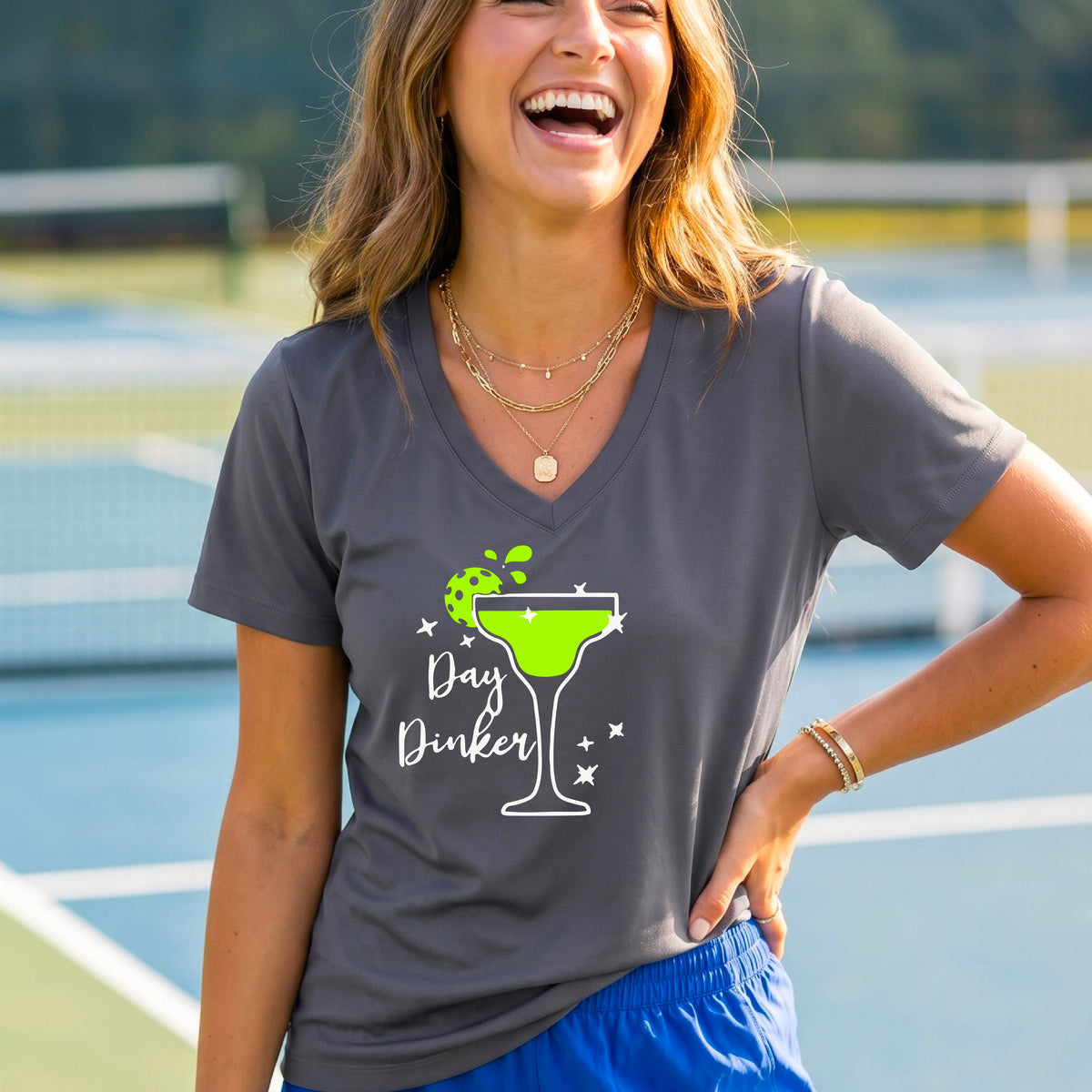 A woman wearing a gray V-neck t-shirt with a 'Day Dinker' pickleball lime garnish martini graphic, standing on a blue pickleball court.