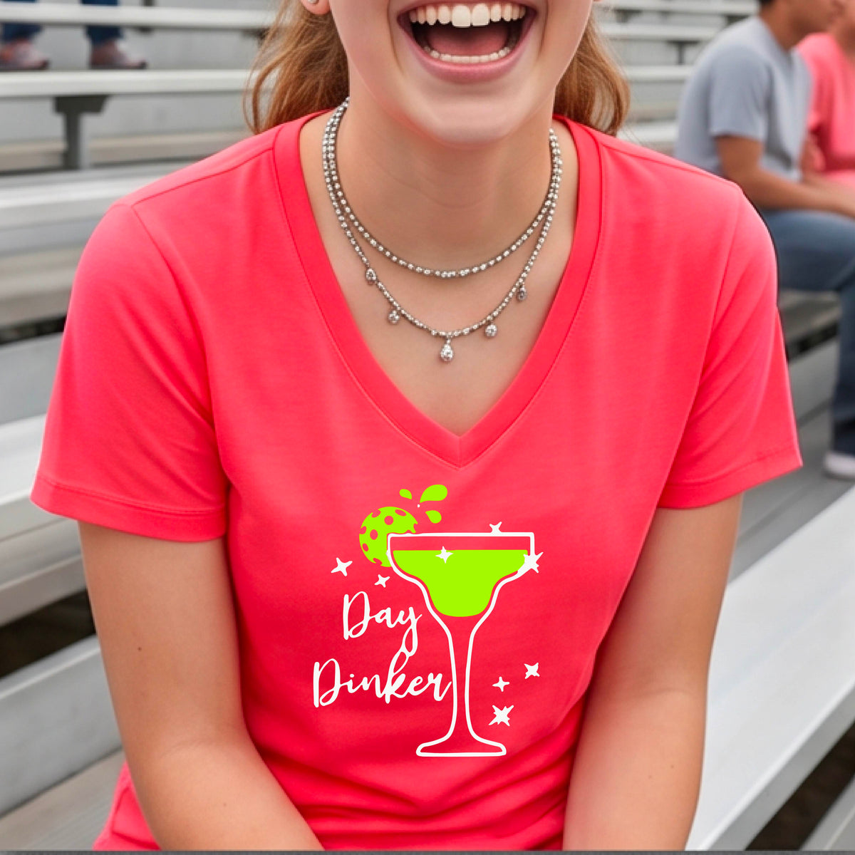 A woman wearing a bright coral pink V-neck t-shirt with a 'Day Dinker' pickleball lime garnish martini graphic, standing on a blue pickleball court.