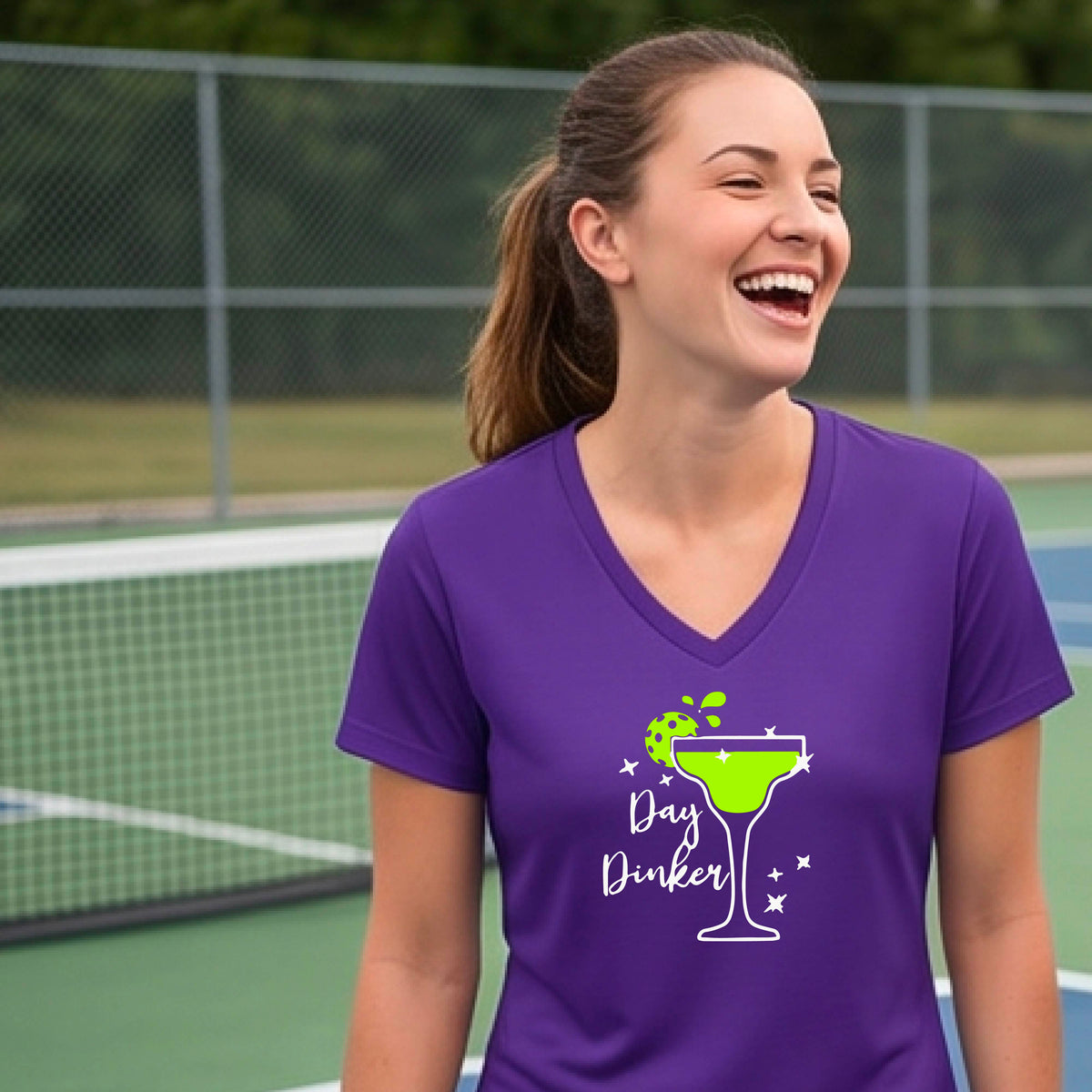 A woman wearing a purple V-neck t-shirt with a 'Day Dinker' pickleball lime garnish martini graphic, standing on a blue pickleball court. 