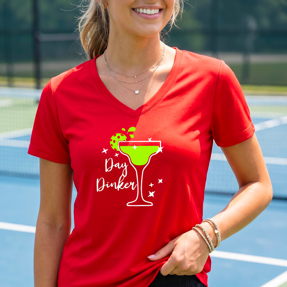 A woman wearing a bright red V-neck t-shirt with a 'Day Dinker' pickleball lime garnish martini graphic, standing on a blue pickleball court.