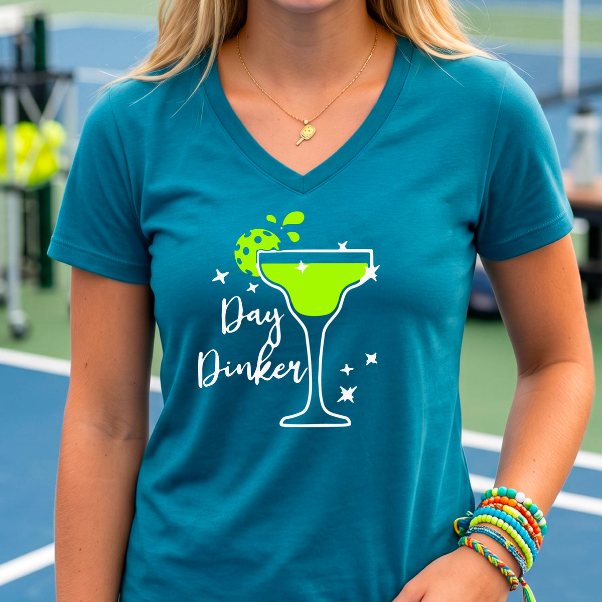 A woman wearing a teal V-neck t-shirt with a 'Day Dinker' pickleball lime garnish martini graphic, standing on a blue pickleball court.