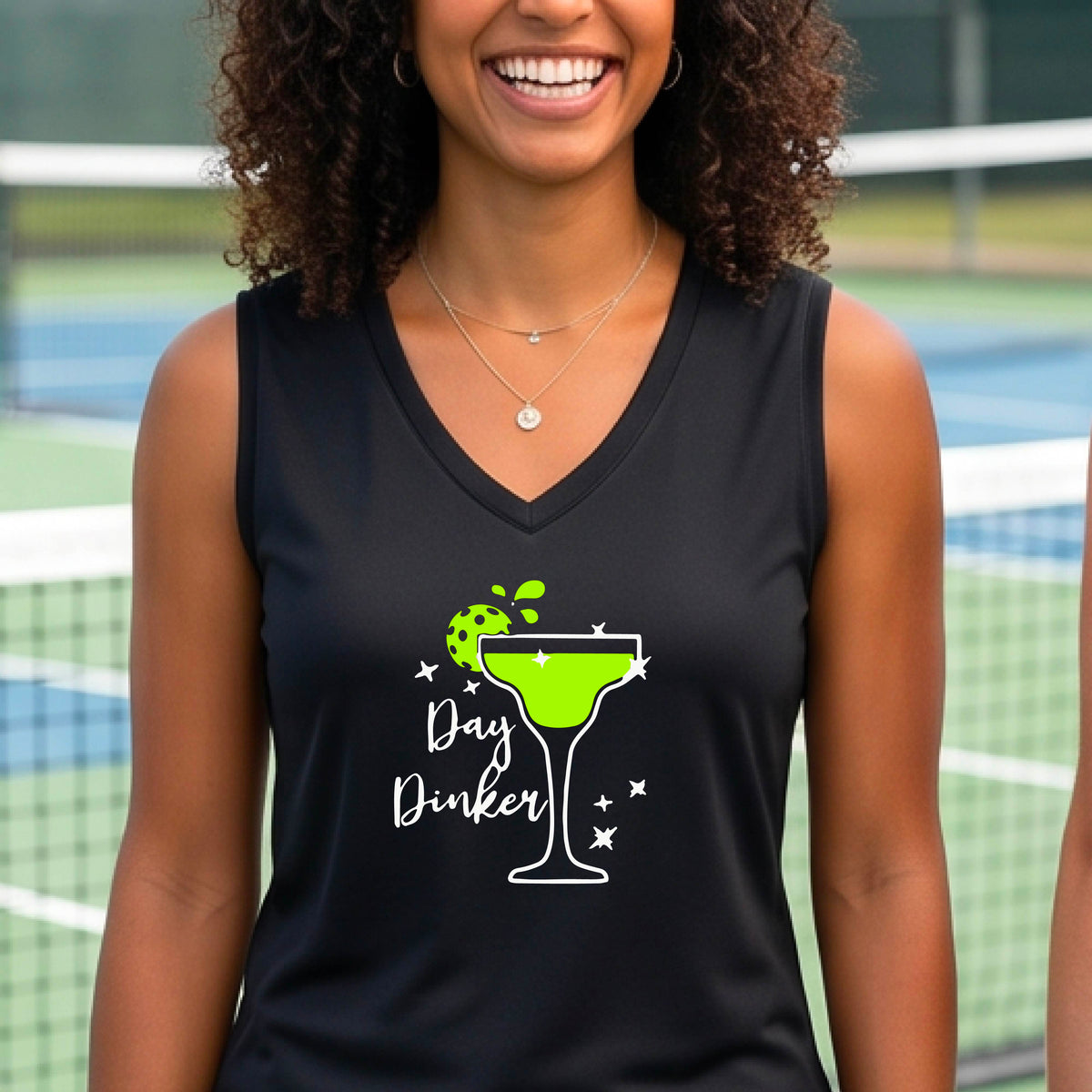 A smiling woman wearing a black sleeveless V-neck tank top with a green cocktail glass and pickleball lime garnish and white 'Day Dinker' text graphic, standing on a pickleall court.