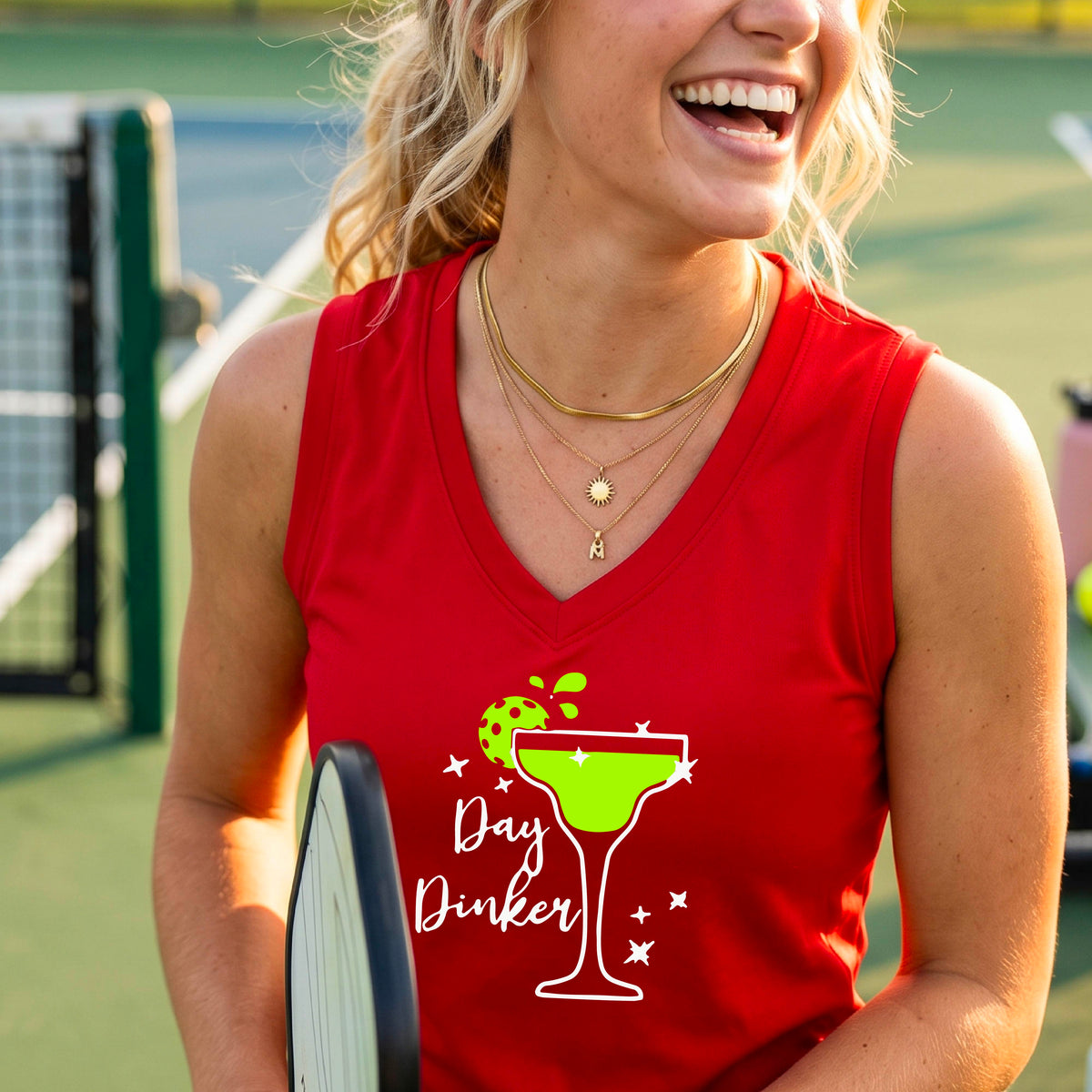 A smiling woman wearing a red sleeveless V-neck tank top with a green cocktail glass and pickleball lime garnish and white 'Day Dinker' text graphic, standing on a pickleall court.
