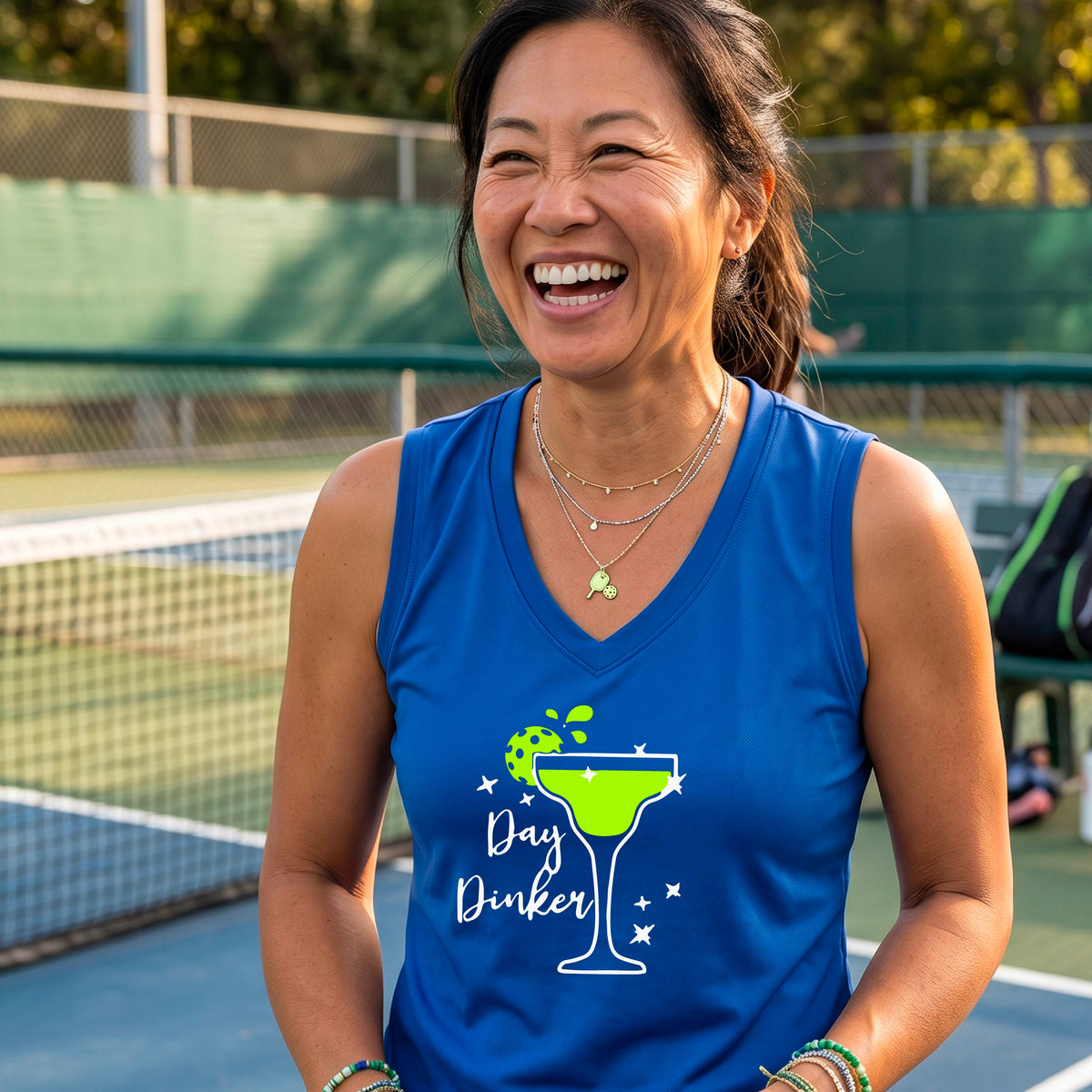 A smiling woman wearing a royal blue sleeveless V-neck tank top with a green cocktail glass and pickleball lime garnish and white 'Day Dinker' text graphic, standing on a pickleall court.