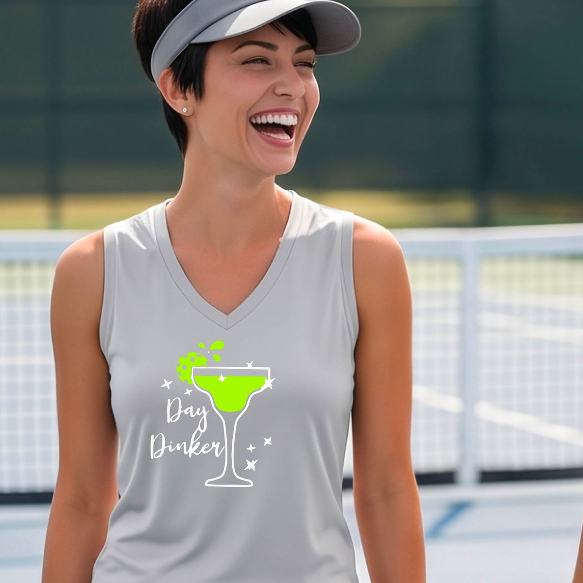A smiling woman wearing a light gray silver sleeveless V-neck tank top with a green cocktail glass and pickleball lime garnish and white 'Day Dinker' text graphic, standing on a pickleall court.