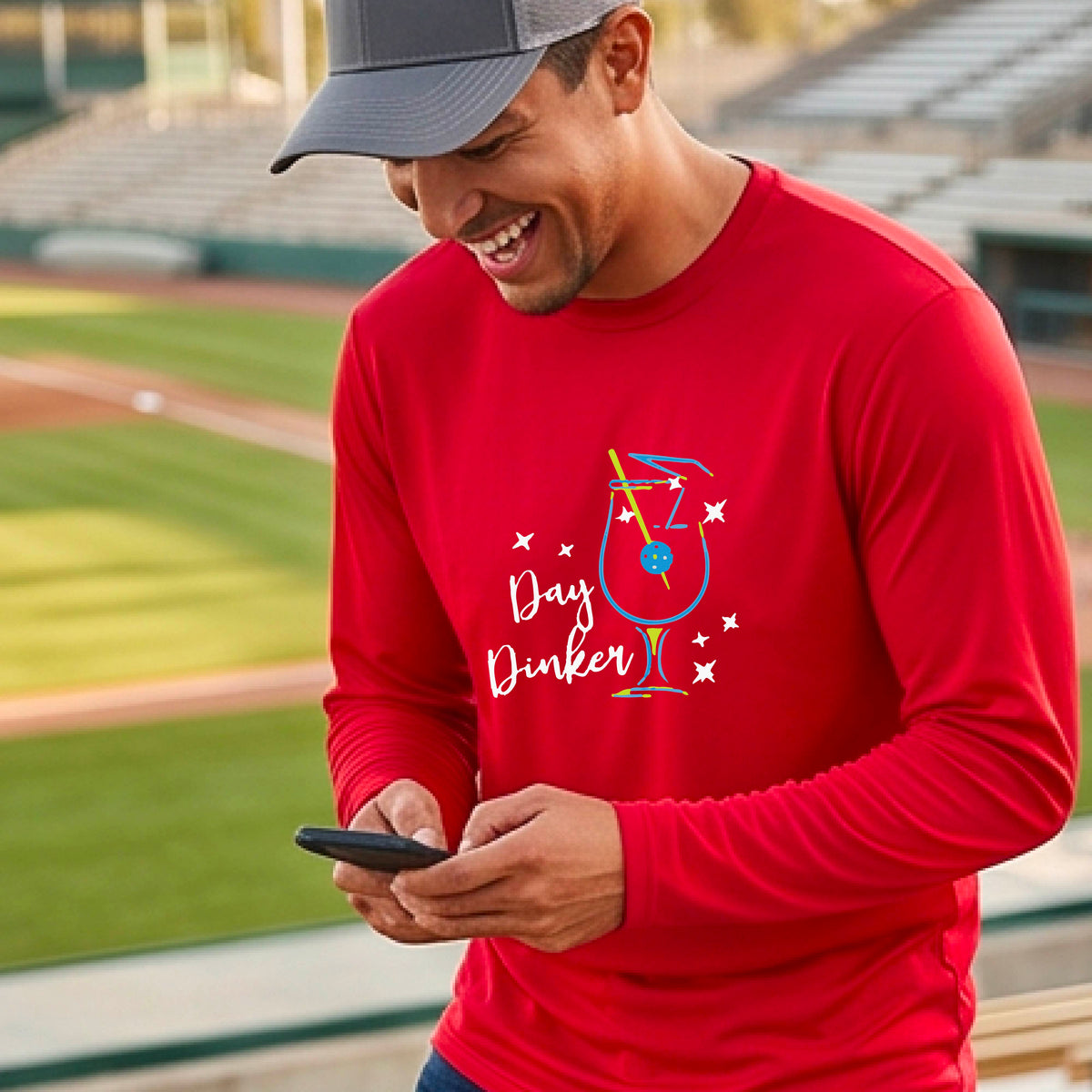 A smiling man wearing a red long-sleeve t-shirt with a 'Day Dinker' cocktail with a pickleall olive garnish graphic, standing on an outdoor pickleball court.
