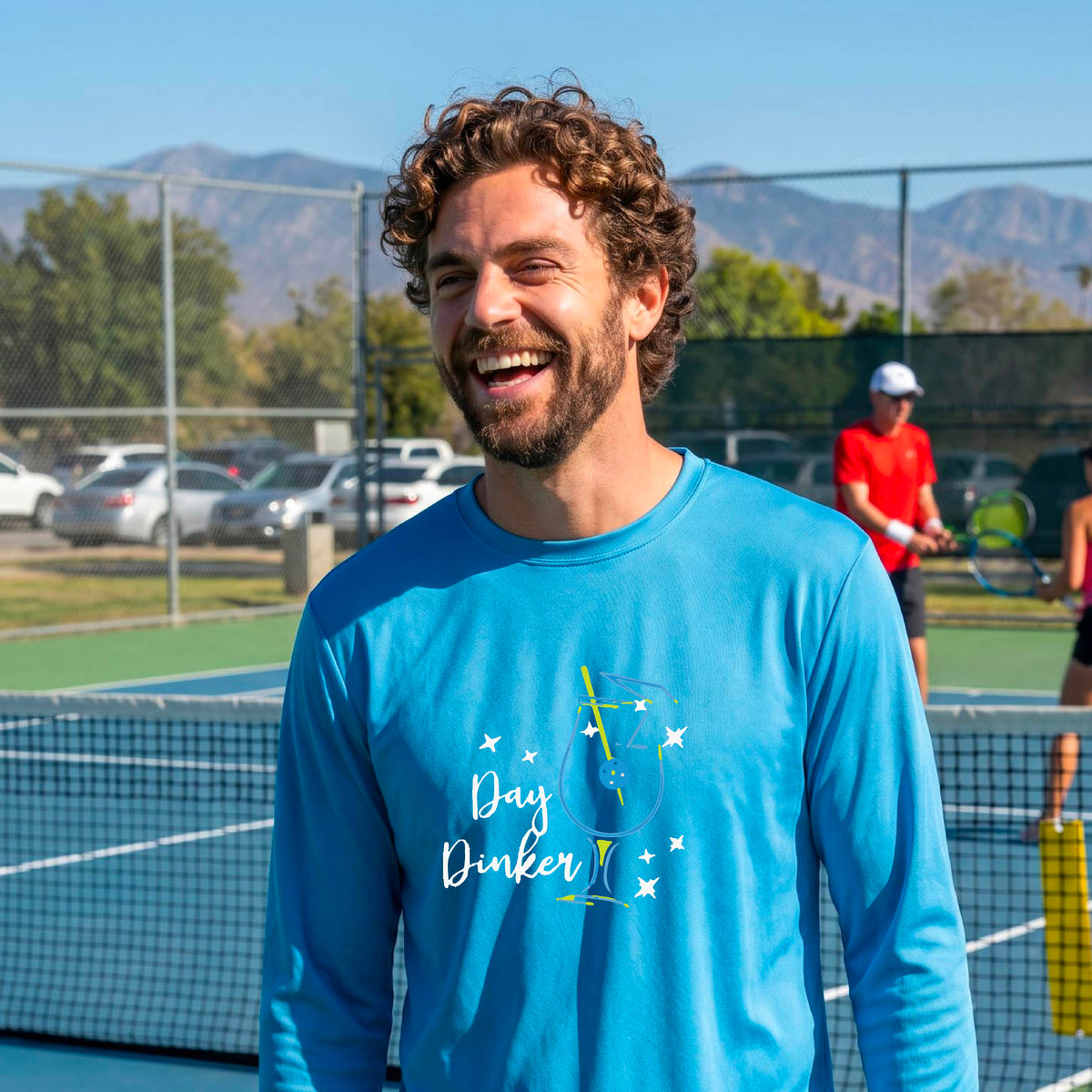A smiling man wearing a light blue long-sleeve t-shirt with a 'Day Dinker' cocktail with a pickleall olive garnish graphic, standing on an outdoor pickleball court.