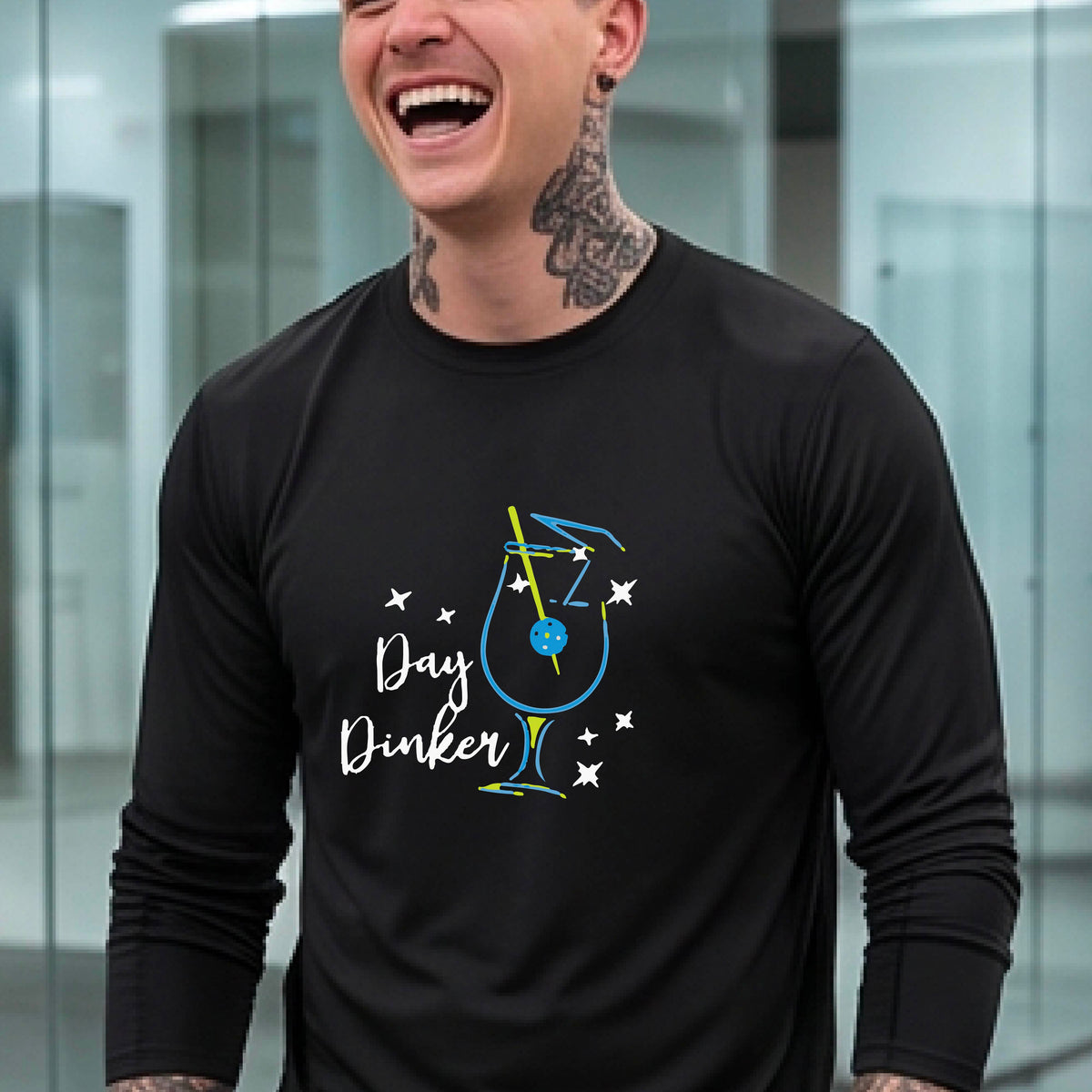 A smiling man wearing a black long-sleeve t-shirt with a 'Day Dinker' cocktail with a pickleall olive garnish graphic, standing on an outdoor pickleball court.