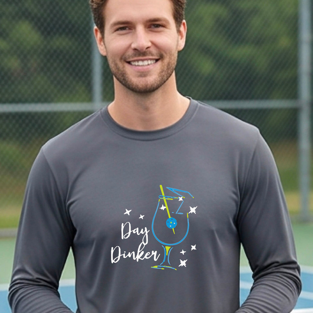 A smiling man wearing a dark gray long-sleeve t-shirt with a 'Day Dinker' cocktail with a pickleall olive garnish graphic, standing on an outdoor pickleball court.