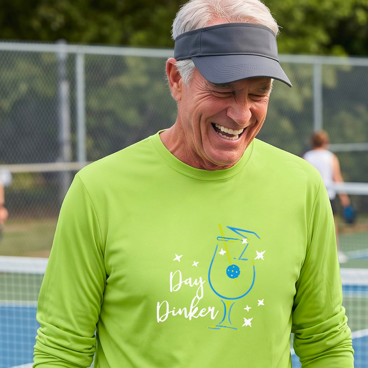 A smiling man wearing a lime green long-sleeve t-shirt with a 'Day Dinker' cocktail with a pickleall olive garnish graphic, standing on an outdoor pickleball court.