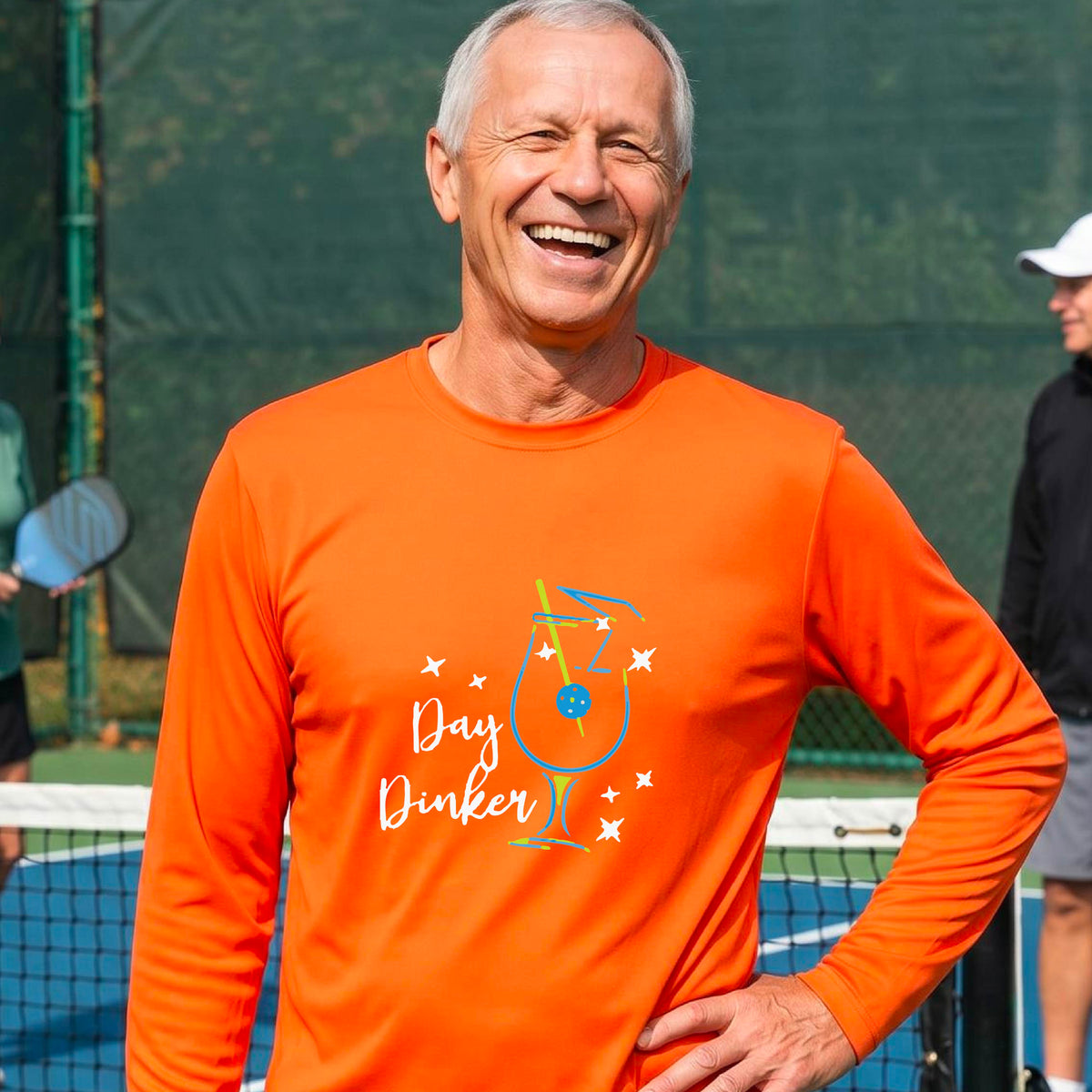 A smiling man wearing an orange long-sleeve t-shirt with a 'Day Dinker' cocktail with a pickleall olive garnish graphic, standing on an outdoor pickleball court.