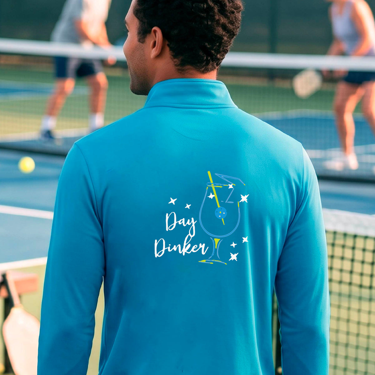 A man viewed from behind wearing a light blue long-sleeve quarter-zip athletic jacket on a pickleball court. The back of the jacket features a colorful graphic of a cocktail martini glass with a blue pickleall olive garnish on it, stars, and the words 'Day Dinker' in white script.