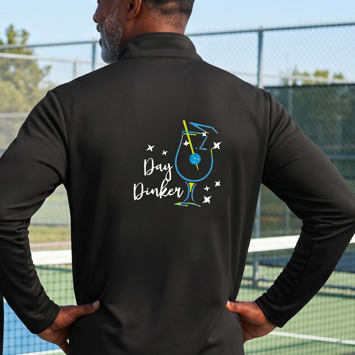 A man viewed from behind wearing a black long-sleeve quarter-zip athletic jacket on a pickleball court. The back of the jacket features a colorful graphic of a cocktail martini glass with a blue pickleall olive garnish on it, stars, and the words 'Day Dinker' in white script.