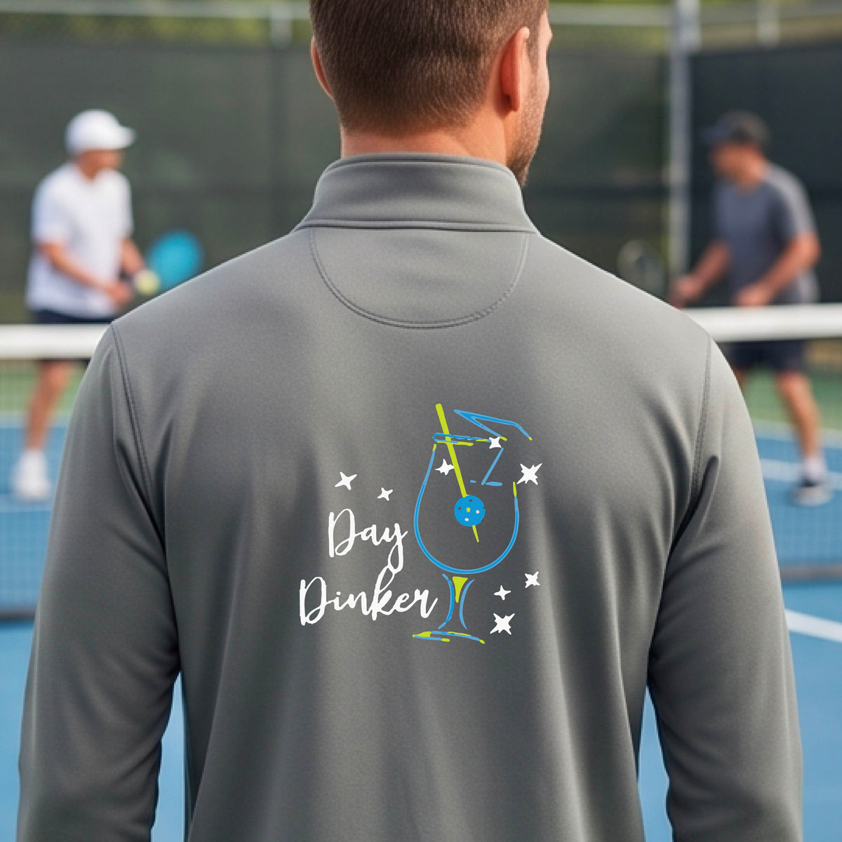 A man viewed from behind wearing a gray long-sleeve quarter-zip athletic jacket on a pickleball court. The back of the jacket features a colorful graphic of a cocktail martini glass with a blue pickleall olive garnish on it, stars, and the words 'Day Dinker' in white script.