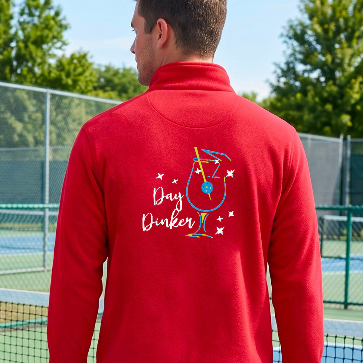 A man viewed from behind wearing a red long-sleeve quarter-zip athletic jacket on a pickleball court. The back of the jacket features a colorful graphic of a cocktail martini glass with a blue pickleall olive garnish on it, stars, and the words 'Day Dinker' in white script.
