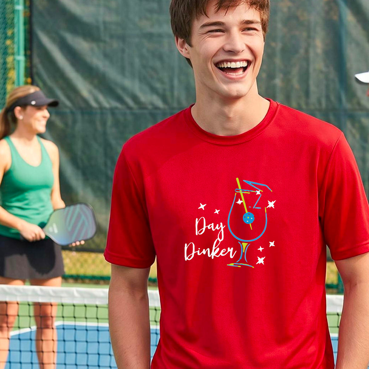 A smiling man wearing a red t-shirt with a 'Day Dinker' cocktail graphic with a pickleball olive garnish stands on an outdoor pickleball court. 