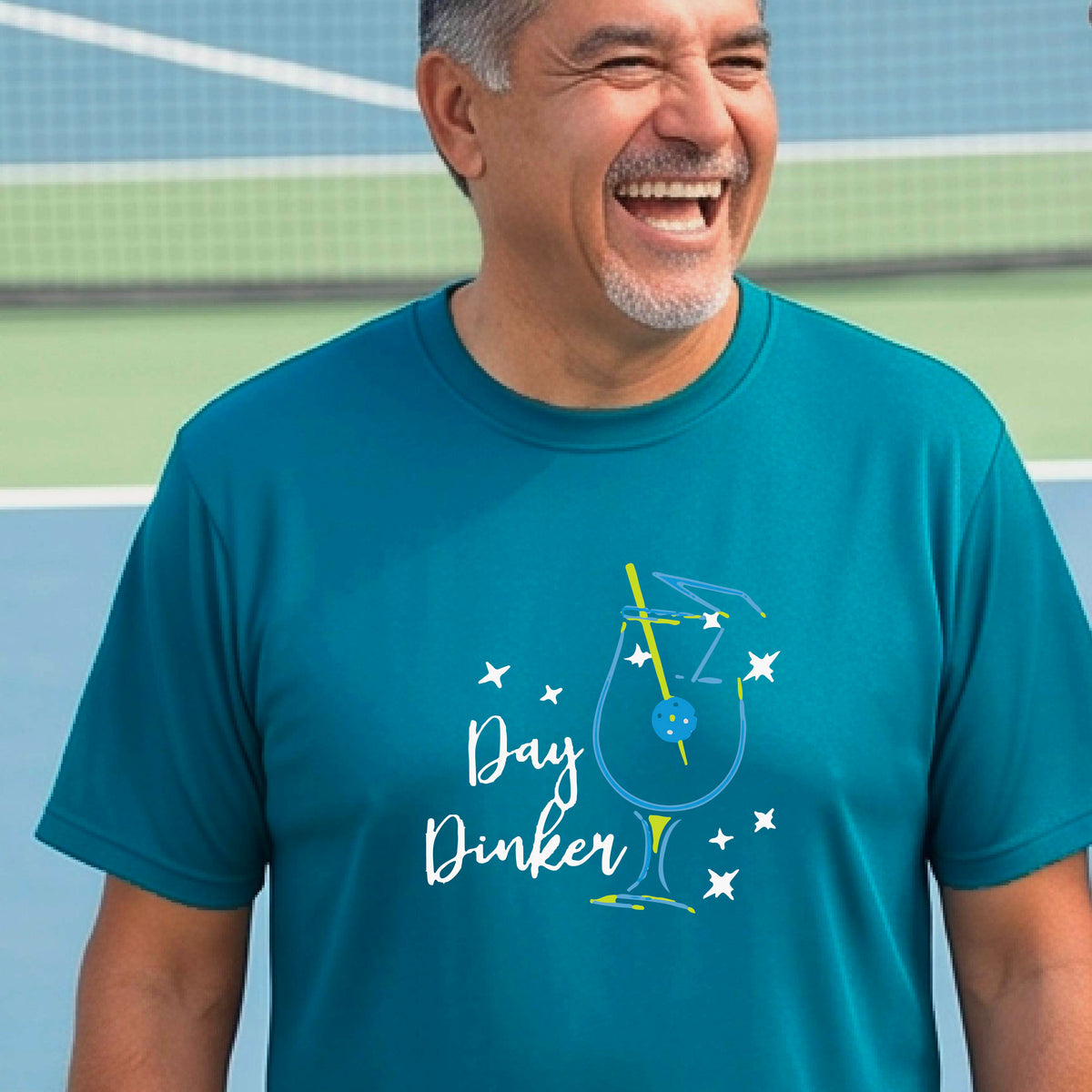 A smiling man wearing a teal t-shirt with a 'Day Dinker' cocktail graphic with a pickleball olive garnish stands on an outdoor pickleball court. 