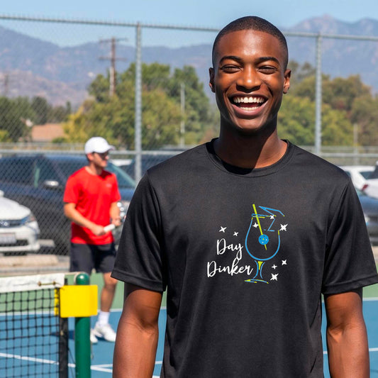 A smiling man wearing a black t-shirt with a 'Day Dinker' cocktail graphic with a pickleball olive garnish stands on an outdoor pickleball court. 