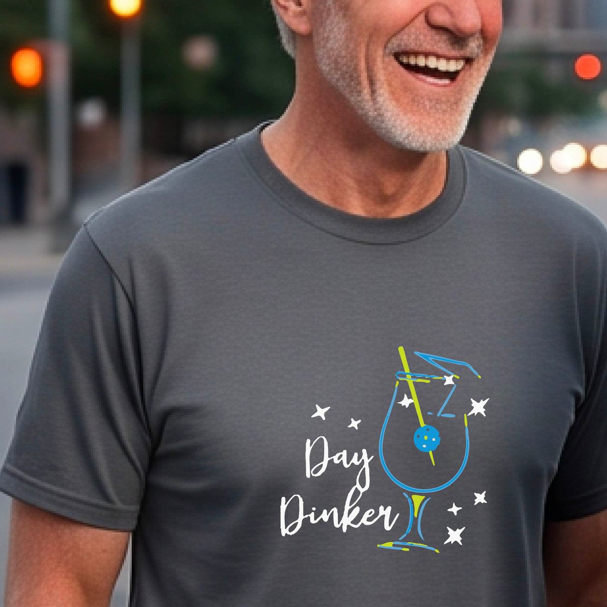 A smiling man wearing a gray t-shirt with a 'Day Dinker' cocktail graphic with a pickleball olive garnish stands on an outdoor pickleball court. 