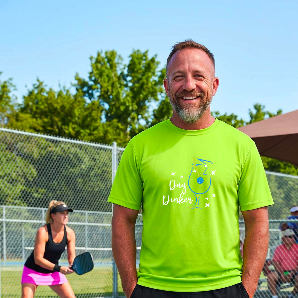 A smiling man wearing a lime green t-shirt with a 'Day Dinker' cocktail graphic with a pickleball olive garnish stands on an outdoor pickleball court. 