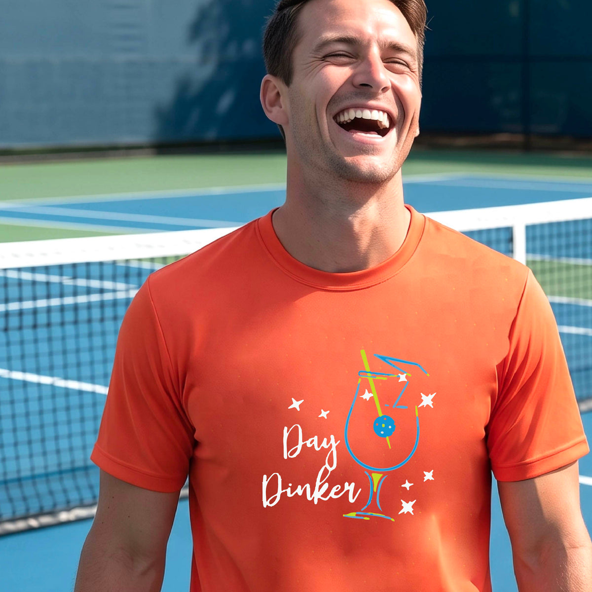A smiling man wearing an orange t-shirt with a 'Day Dinker' cocktail graphic with a pickleball olive garnish stands on an outdoor pickleball court. 