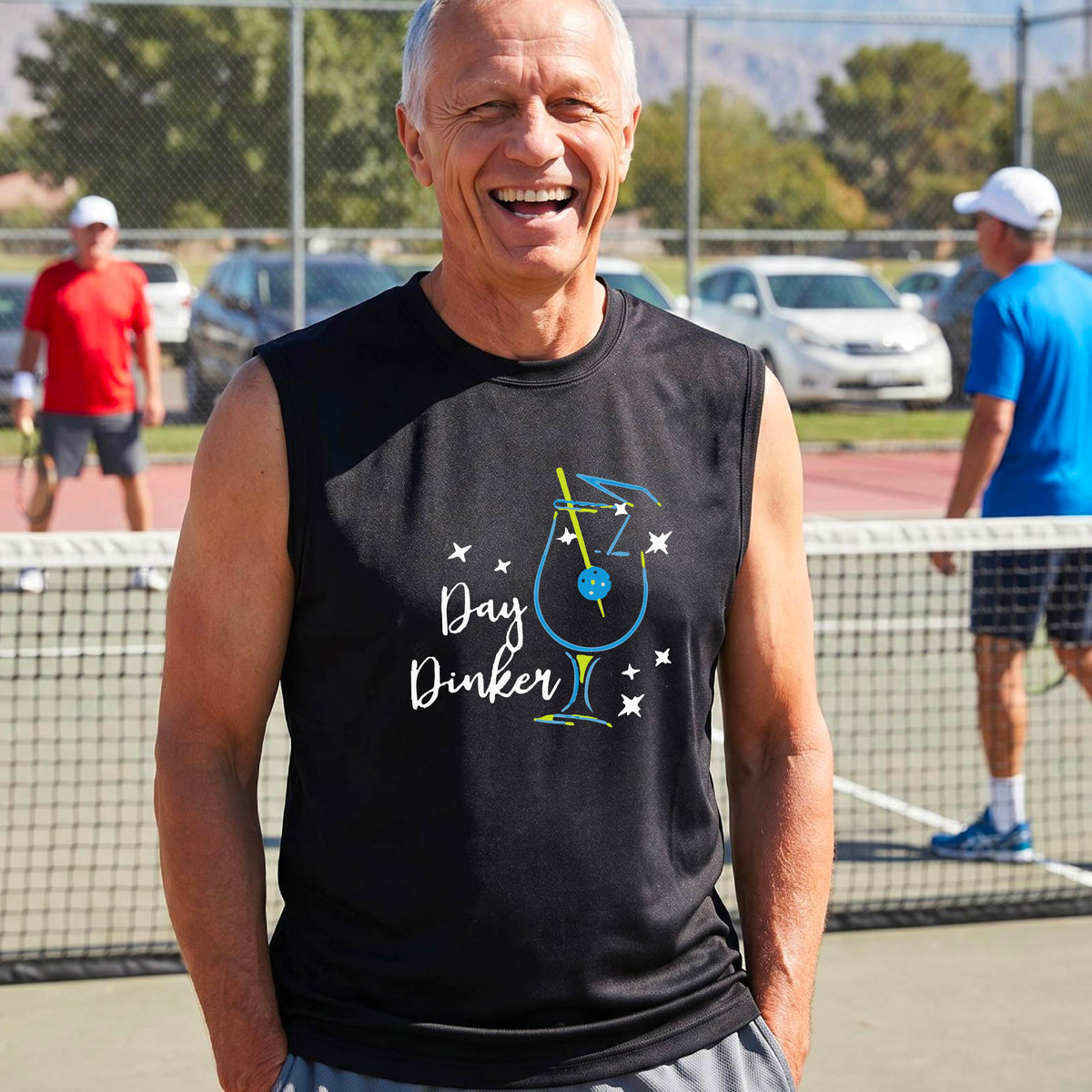 A smiling older man wearing a black sleeveless graphic t-shirt stands on a tennis court. The shirt features a colorful cocktail martini glass with a pickleball olive garnish sign with the words 'Day Dinker' in white script and small white stars.