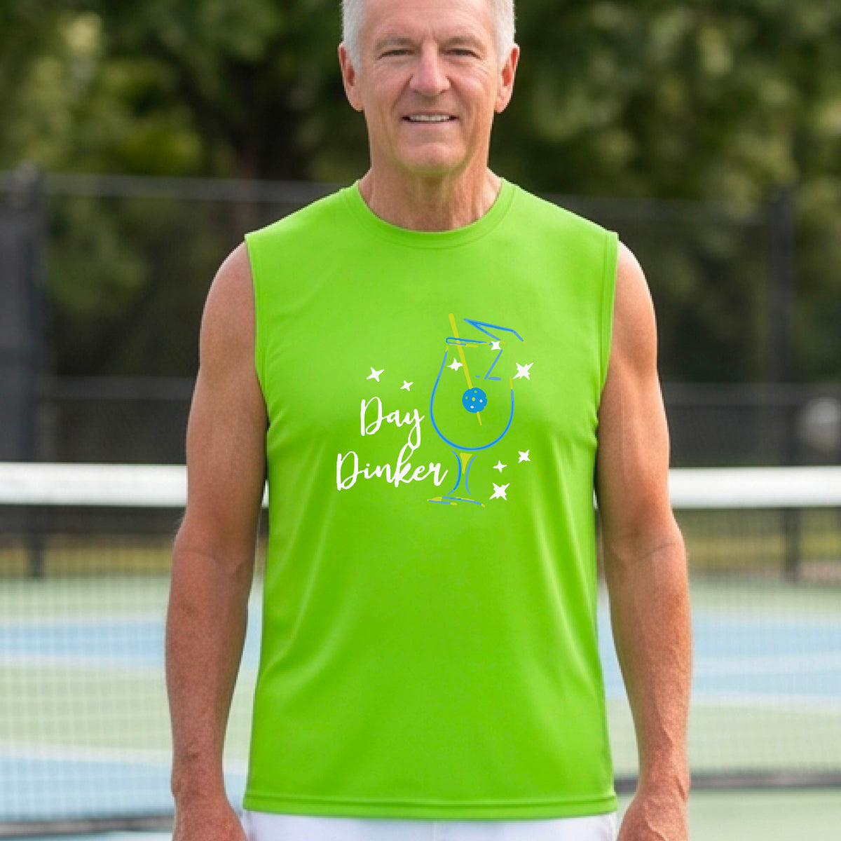 A smiling older man wearing a lime green sleeveless graphic t-shirt stands on a tennis court. The shirt features a colorful cocktail martini glass with a pickleball olive garnish sign with the words 'Day Dinker' in white script and small white stars.