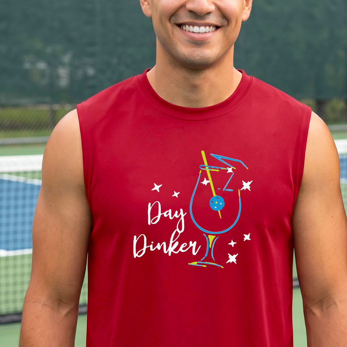 A smiling older man wearing a red sleeveless graphic t-shirt stands on a tennis court. The shirt features a colorful cocktail martini glass with a pickleball olive garnish sign with the words 'Day Dinker' in white script and small white stars.