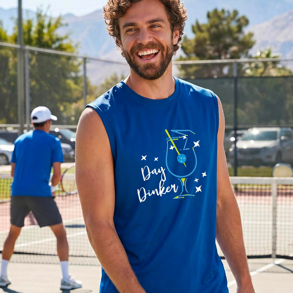 A smiling older man wearing a royal blue sleeveless graphic t-shirt stands on a tennis court. The shirt features a colorful cocktail martini glass with a pickleball olive garnish sign with the words 'Day Dinker' in white script and small white stars.