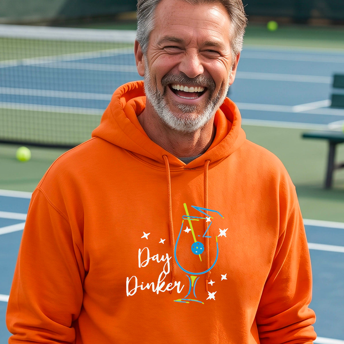 A man wearing an orange hoodie with a colorful graphic on the chest that reads 'Day Dinker' alongside a martini cocktail glass with a pickelball olive garnish illustration.